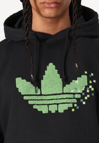 Svart hoodtröja med en texturerad grön Adidas-logotyp i pixelmönster med varierande nyanser av grönt och blått i nedre högra hörnet.
