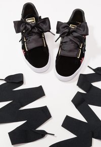Baskets Puma en velours noir avec larges lacets en ruban de satin noués en nœuds, broderies florales sur les côtés et semelles en caoutchouc blanc.