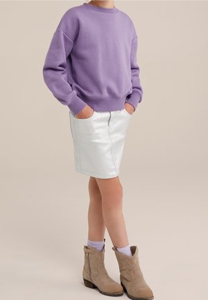 Kind draagt een paarse trui, witte rok, paarse sokken en beige enkellaarsjes, staat met handen in de zakken tegen een neutrale achtergrond.