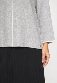 Main d'une femme à côté d'un haut gris clair à manches longues avec bordure blanche et jupe plissée noire sur fond blanc.