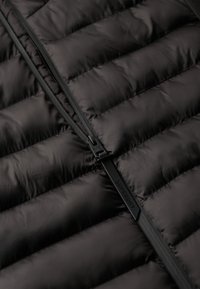 Sort puffer vest med quiltede sektioner og en central lynlås med mærke-træk. Materialet ser skinnende og letvægts ud.