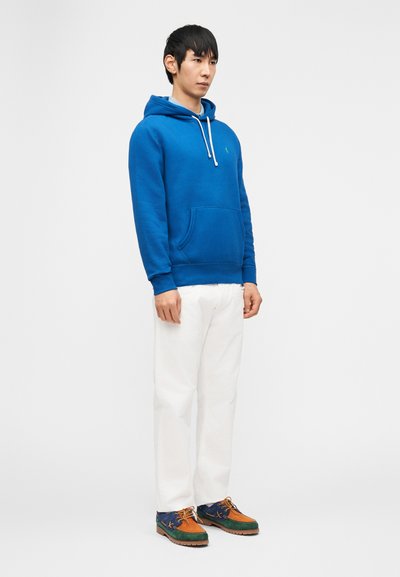 Άνδρας που στέκεται φορώντας ένα μπλε φούτερ με κουκούλα, λευκό παντελόνι και πολύχρωμα αθλητικά παπούτσια μπροστά σε απλό φωτεινό φόντο.