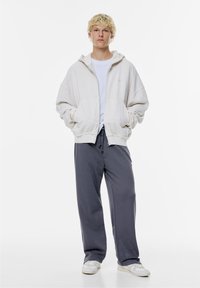 Licht crème zip-up hoodie met een trekkoordcapuchon, gecombineerd met donkergrijze joggingbroek. Getextureerde stof en een losse pasvorm kenmerken de kleding.