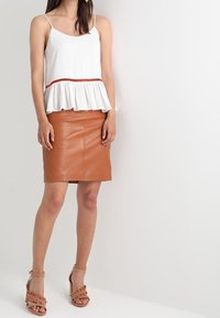 Top bianco senza maniche con orlo arricciato e accentuazione rossa, abbinato a una gonna in pelle marrone aderente e sandali con zeppa color marrone chiaro e fasce.