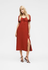 Žena v tehlovočervenom midi šatách s krátkymi rukávmi, so zaväzovaním vpredu, vysokým rozparkom na stehne a čiernymi sandálmi na vysokom opätku so zaväzovaním.