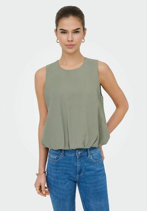 Femme aux cheveux attachés portant un haut sans manches vert sauge et un jean bleu, debout avec un bras derrière le dos.