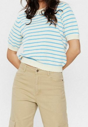 Woman wearing a white and light blue striped short-sleeve knit top with scalloped edges and beige high-waisted pants with pockets.
