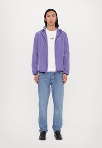 Casaco leve lilás com capuz, usado sobre uma t-shirt branca com um logótipo, combinado com jeans azuis e sapatos pretos. Design simples, ajuste casual.