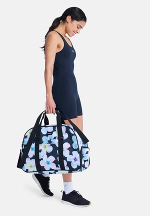 Jonge vrouw in zwarte sportkleding die een zwarte duffeltas met bloemmotief vasthoudt en met een lichte glimlach loopt tegen een effen achtergrond.