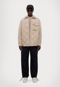 Giacca trapuntata beige con tasca sul petto, indossata sopra un maglione chiaro, abbinata a pantaloni neri e sneakers bianche. Cappellino nero e occhiali da sole inclusi.