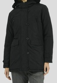 Svart, knäppt vinterjacka med framfickor och blixtlåsfickor på bröstet, bärs över rutiga byxor.