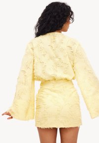 Robe mini jaune avec un tissu texturé et effiloché, à manches cloche longues, mettant en avant une taille cintrée et un dos droit.