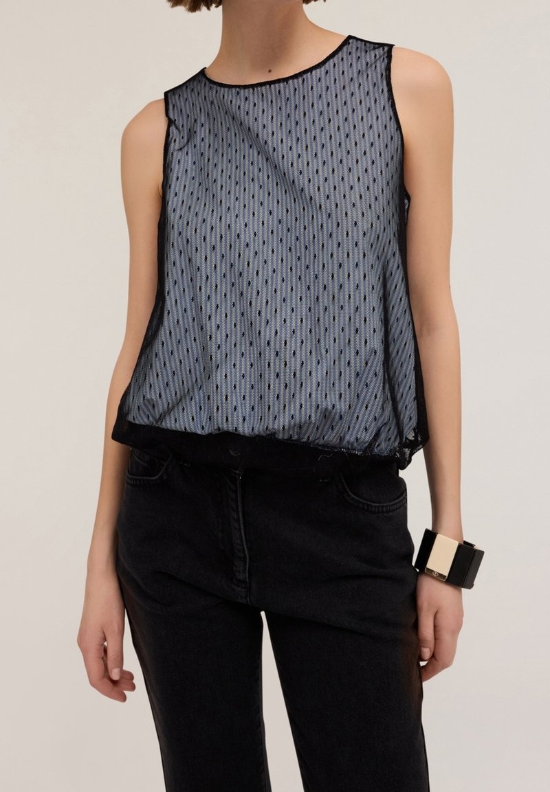 Femme portant un haut noir transparent sans manches à pois par-dessus un débardeur gris, un pantalon noir et un bracelet épais noir et blanc au poignet droit.