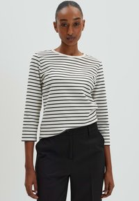 Camicia a maniche lunghe a righe con righe orizzontali nere e bianche, scollo rotondo e tessuto morbido. Indossata con pantaloni neri su misura.