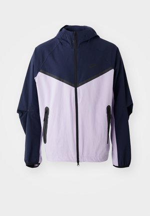 Nike lettvektsjakke, marineblå og lyse lilla fargeblokker, glidelås foran, hette, har svarte glidelåser og halvpaneldesign på skuldrene.