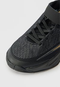 Črne športne čevlje z teksturiranim mrežastim zgornjim delom, ravnimi vezalkami in gumijastim podplatom. Opremljeni so z ojačanim zaščitnim delom na prstih in diskretnimi zlatenimi detajli.