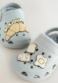 Gray clogs with fuzzy interior feature food-themed rubber charms: croissant, bread, milk carton, and egg. Ventilation holes are present.
