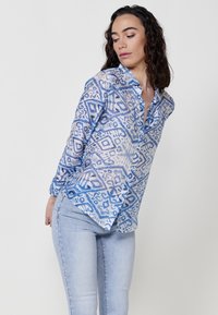 Blusa leggera e trasparente di colore blu con motivi geometrici, chiusura con bottoni e maniche lunghe, abbinata a jeans attillati di un blu chiaro.