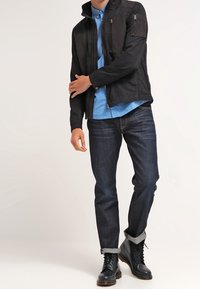 Giacca nera, leggera, con tasche con zip, abbinata a una camicia a quadri blu, jeans scuri e stivali neri stringati.