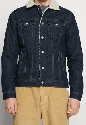 Mand iført en mørkeblå denimjakke med hvidt lammeskindskrage og beige bukser i kordfløjl, stående foran en ensfarvet baggrund.