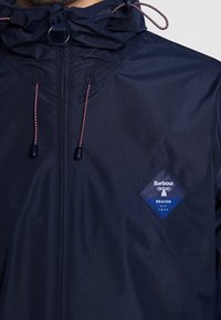 Granatowa wodoodporna kurtka z kapturem na sznurek, ozdobiona niebieską naszywką z logo w kształcie diamentu oraz kontrastowymi akcentami ze sznurka przy zamku.