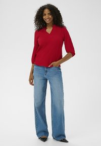 Rød ribbet pullover med åbning ved halsen, trekvartlange ærmer, parret med lyseblå vide jeans og sorte spidse sko.