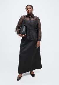 Γυναίκα με διάφανη μαύρη μπλούζα πάνω από μαύρο κορμάκι και μακριά μαύρη φούστα, κρατάει μαύρη τσάντα, στέκεται σε λευκό φόντο.