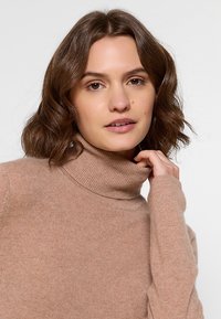 Beige Rollkragenpullover aus weichem, strukturiertem Stoff mit langen Ärmeln. Das Design verfügt über einen eng anliegenden Kragen und eine lässige Silhouette.
