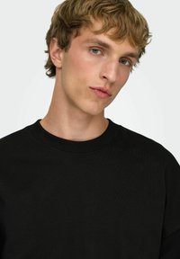 Sudadera negra de algodón con cuello redondo acanalado y tejido texturizado. Vista en primer plano del rostro, con cabello claro y ojos azules.