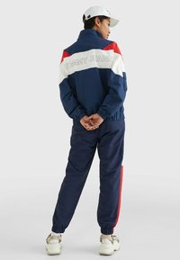 Marineblå vindjakke med røde og hvide detaljer, elastiske manchetter og "TOMMY JEANS" logo på ryggen, parret med matchende bukser og sneakers.