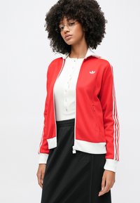 Jauna sieviete ar cirtainiem matiem, valkā brilles, sarkanu Adidas jaku ar baltām svītrām, baltu pogājamu kreklu un melnu svārku.