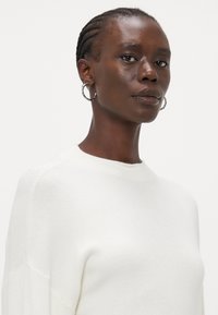 Pull en tricot blanc avec un col rond, arborant un motif texturé. Le modèle a les cheveux courts tressés et porte des boucles d'oreilles en argent.