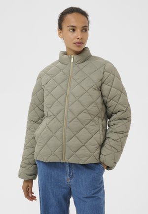 Frau, die eine hellolivengrüne gesteppte Jacke mit goldenen Reißverschluss und eine blaue Jeans trägt, mit einer Hand in der Tasche steht und seitlich schaut.