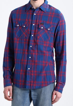 Homme portant une chemise à carreaux bleu et rouge à manches longues avec des poches poitrine à rabat et un jean bleu clair.