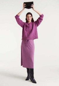 Jersey de punto texturizado morado, falda midi morada a juego con una abertura lateral, botas negras hasta la rodilla y un bolso negro sostenido sobre la cabeza.