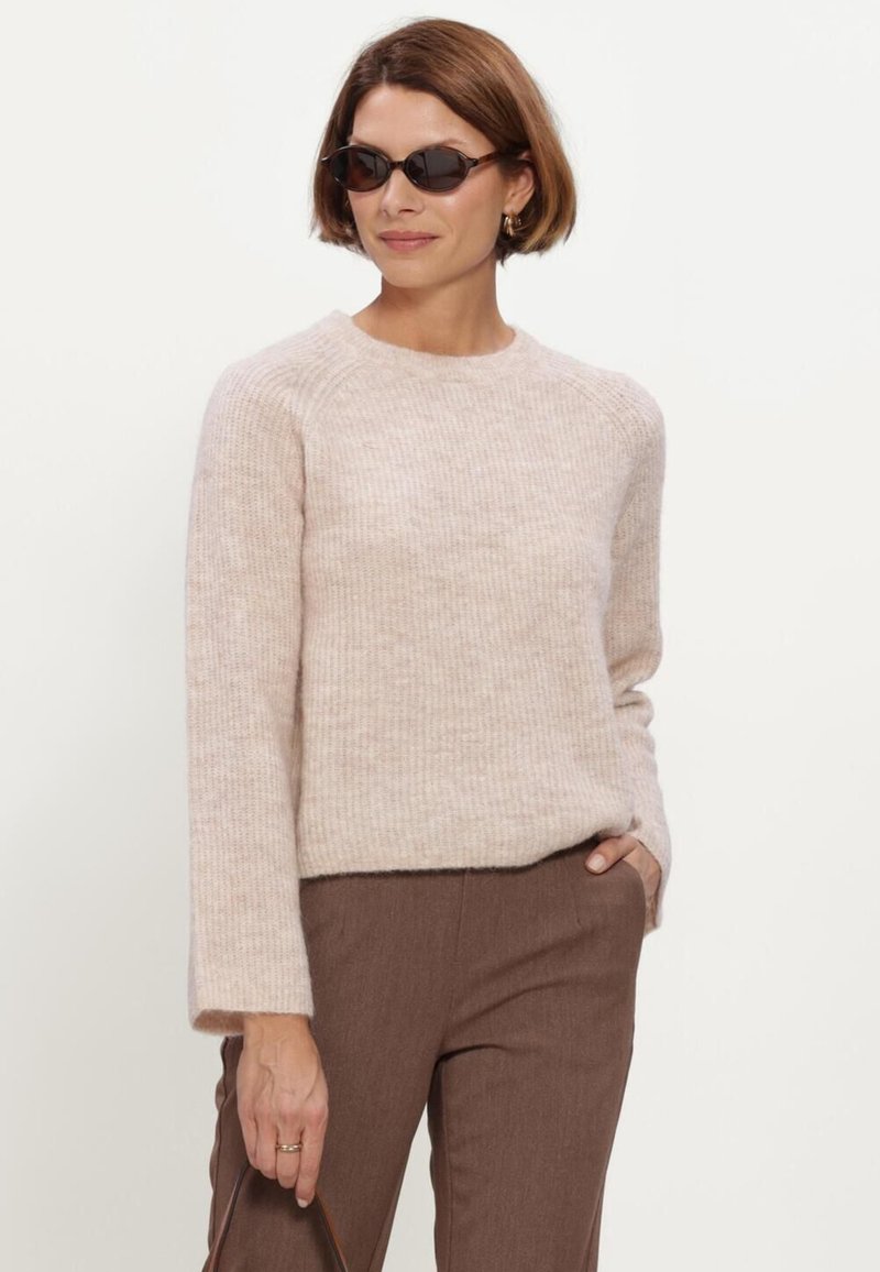 Femme portant des lunettes de soleil foncées, un pull en tricot beige clair, un pantalon marron et tenant un sac à main sur un fond blanc.