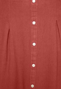 Tissu couleur rouille avec une rangée verticale de sept boutons blancs et des plis cousus de chaque côté.