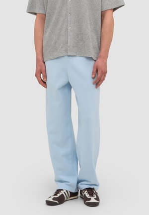 Persoon draagt lichtblauwe losse broek, gestructureerd grijs T-shirt met korte mouwen en bruine en witte sneakers, staand tegen een effen achtergrond.