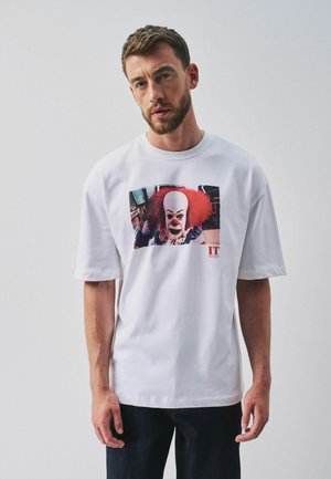 Man with short brown hair and beard wearing white T-shirt with a colorful clown face graphic from the movie "IT" on a plain background.