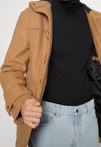 Wollmischungsjacke in Camel-Farbe mit Kapuze, hölzernen Toggle-Knöpfen und einem schwarzen gerippten Rollkragenpulli darunter; kombiniert mit hellblauen Jeans.