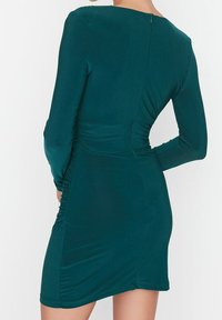 Robe ajustée à manches longues en teal, fabriquée dans un tissu doux. Présente des détails froncés sur les côtés et une fermeture éclair discrète au dos.