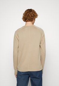 Mężczyzna z kręconymi włosami, ubrany w beżową koszulę z długim rękawem i niebieskie spodnie, odwrócony plecami na tle jasnobiałym.