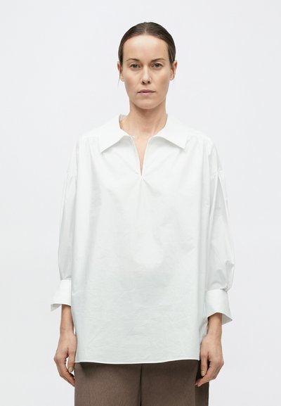 Hátrafogott hajú nő, laza, széles galléros fehér hosszú ujjú blúzt és barna nadrágot viselve, egyszínű fehér háttér előtt áll.