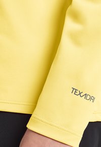 Gelbes kurzärmliges Hemd aus strukturiertem Stoff, mit einem gedruckten "TEXADRI"-Logo am Bündchen. Nahtdetails sind entlang des Ärmels sichtbar.