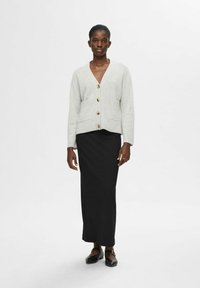 Šviesiai pilkas megztas cardiganas su V formos iškirpte, priekinėmis kišenėmis ir smėlio spalvos mygtukais, derinamas su tvirtai prigludusia juoda maksi sijonu ir plokščiomis bateliais.