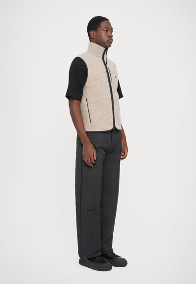 Graue Fleece-Weste mit hohem Kragen, Reißverschluss vorne und Seitentaschen, getragen über einem schwarzen kurzärmligen Shirt und schwarzen gesteppte Hosen.