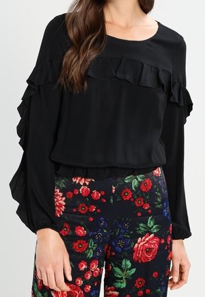 Kobieta w czarnej bluzce z długim rękawem z falbankami oraz w spodniach z wysokim stanem w kolorowy, czerwono-zielony kwiatowy wzór.