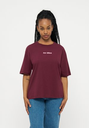 Νεαρή γυναίκα με μακριά πλεξούδα φορούσε μαρουνά t-shirt με την επιγραφή "δεν έχω ιδέα" και μπλε τζιν, στέκεται μπροστά σε λευκό απλό φόντο.