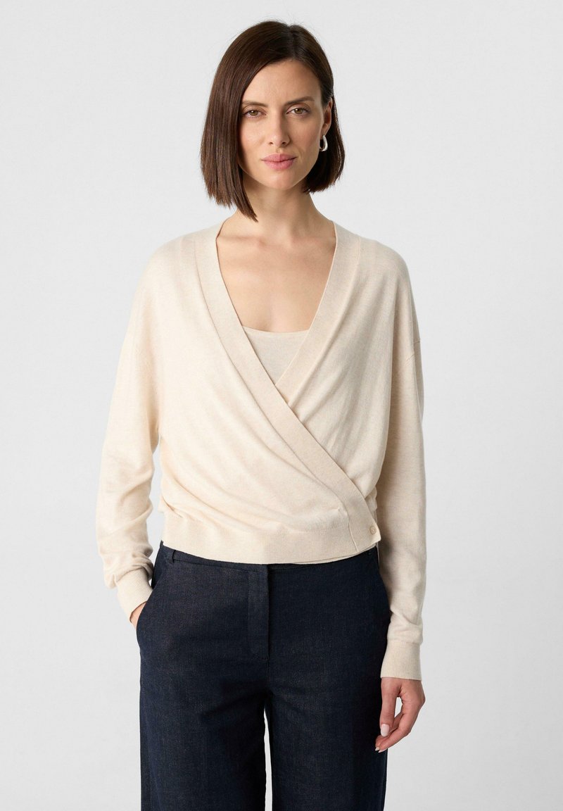 Cardigan beige in tessuto morbido, con scollo a V profondo e maniche lunghe, abbinato a pantaloni scuri e sartoriali. Design minimalista con dettaglio dei bottoni.