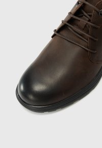 Botine din piele maro, cu un finisaj neted, bot rotund și talpă din cauciuc negru. Prezintă șireturi maronii și un design clasic.